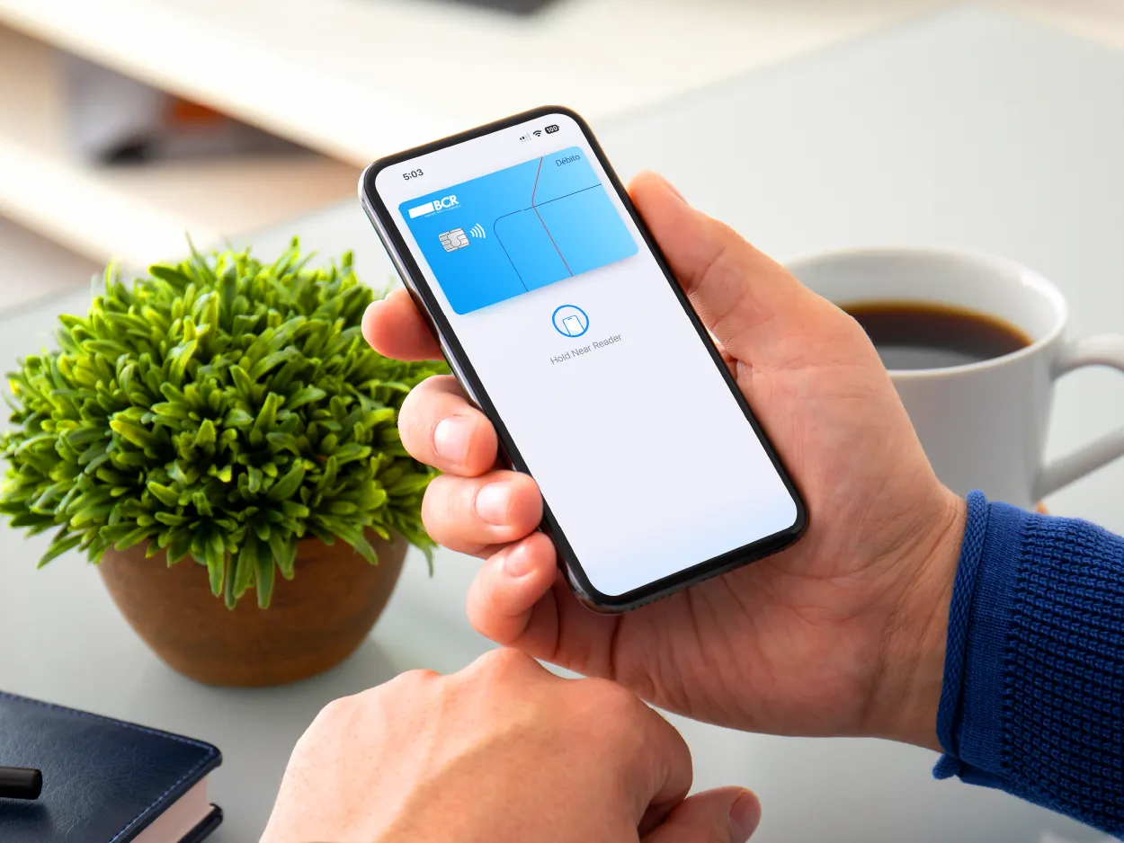Persona sosteniendo un teléfono móvil que muestra una tarjeta digital del BCR en pantalla, sobre una mesa con una planta y una taza de café.