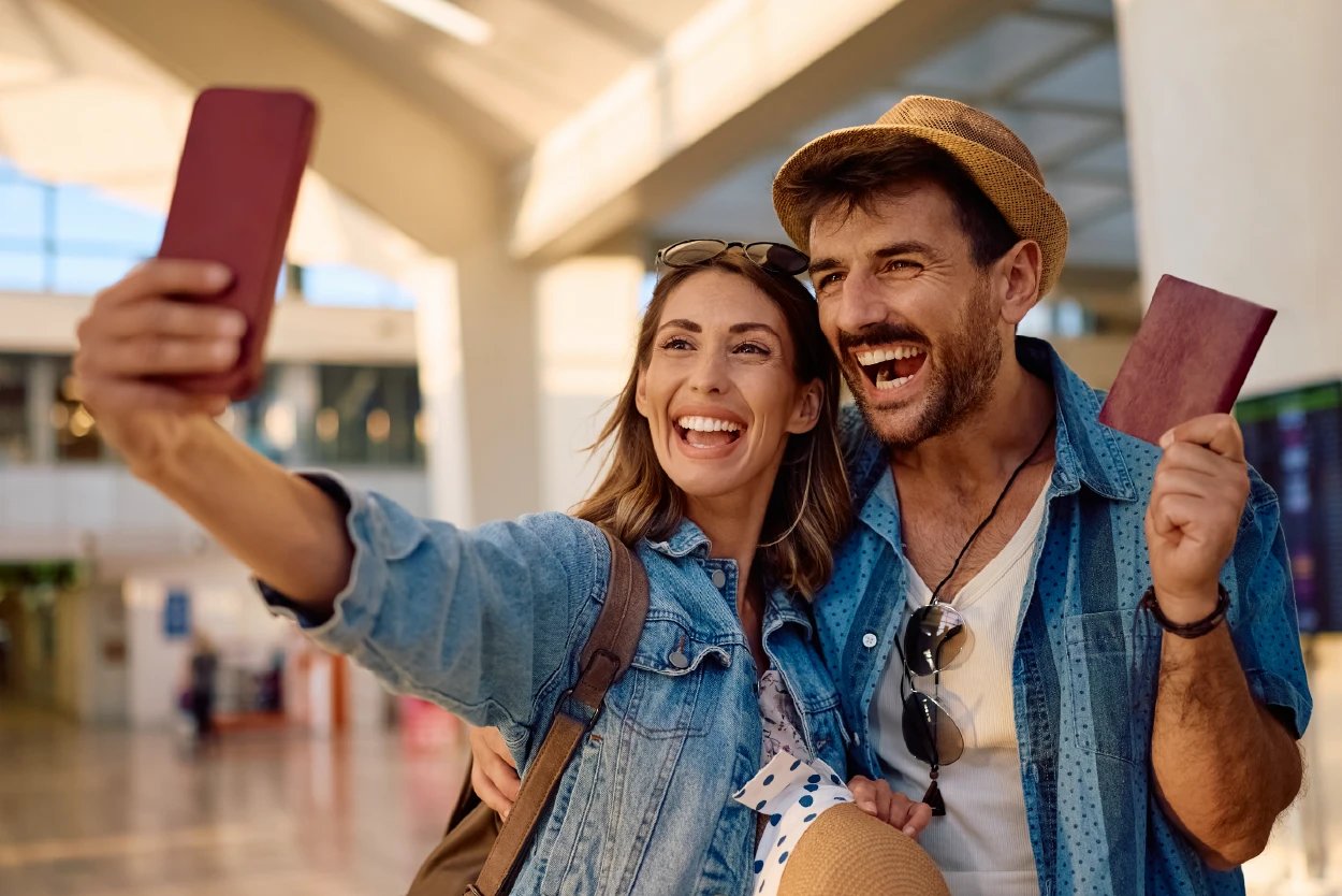 Pareja joven sonriente tomándose una selfie en un aeropuerto, sosteniendo pasaportes y boletos de viaje, mostrando entusiasmo antes de abordar